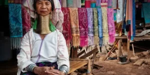 Donna anziana della tribù Kayan (conosciute come Long Neck) con i tradizionali anelli di ottone al collo, seduta in un Hill Tribe Village nel nord della Thailandia con tessuti colorati sullo sfondo.