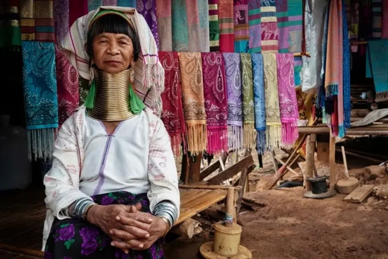 Donna anziana della tribù Kayan del nord Thailanida (conosciute come Long Neck) con i tradizionali anelli di ottone al collo, seduta in un Hill Tribe Village nel nord della Thailandia con tessuti colorati sullo sfondo.