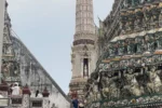 Dettaglio delle sculture e dei mosaici in porcellana sulle terrazze del prang centrale del Wat Arun a Bangkok, con turisti che ammirano l'architettura.