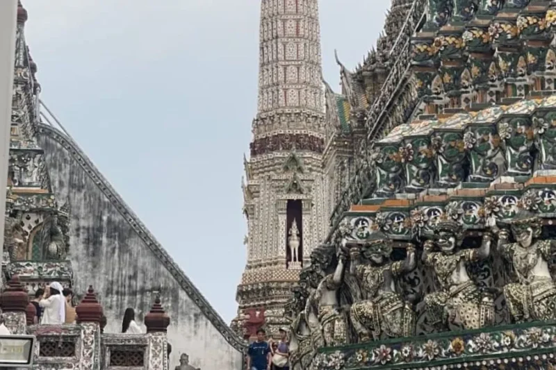 Dettaglio delle sculture e dei mosaici in porcellana sulle terrazze del prang centrale del Wat Arun a Bangkok, con turisti che ammirano l'architettura.