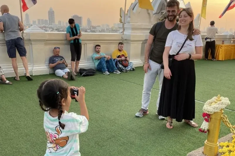 Una bambina thailandese scatta una foto ricordo a due turisti sulla terrazza panoramica del Wat Saket a Bangkok durante il tramonto.