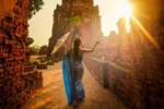 Donna in abito tradizionale e con un ombrello decorativo cammina tra antiche rovine di Ayutthya al tramonto