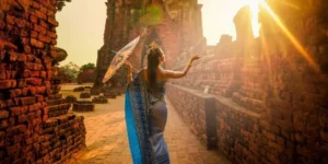 Donna in abito tradizionale e con un ombrello decorativo cammina tra antiche rovine di Ayutthya al tramonto