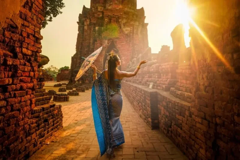 Donna in abito tradizionale e con un ombrello decorativo cammina tra antiche rovine di Ayutthya al tramonto